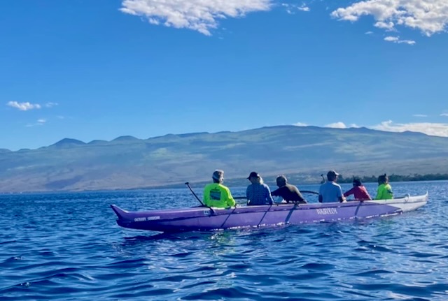A six-person outrigger canoe crew paddling on calm water with mountains behind